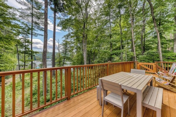 A wooden deck with a table and chairs, surrounded by a wooden railing, overlooking a forested area. - Lakefront home with amazing views, fireplace, firepit, & gas grill (Ludlow)
