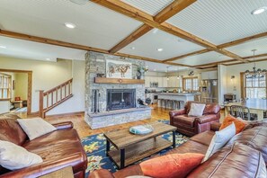 A spacious living room with a stone fireplace, leather sofas, a wooden coffee table, and a high ceiling with exposed beams. - Lakefront home with amazing views, fireplace, firepit, & gas grill (Ludlow)