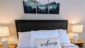 A bed with a 'welcome' pillow, a mountain landscape painting above it, and two bedside lamps on wooden nightstands.