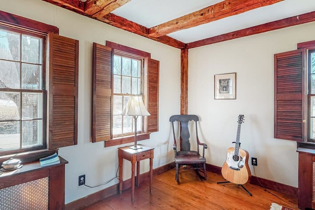 A room with wooden beams, a guitar, a chair, and a table with a lamp.