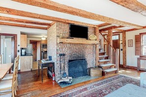 Ein rustikales Interieur mit freiliegenden Holzbalken, einem Kamin aus Backsteinen, einem darüber montierten Fernseher und einer Treppe, die in einen anderen Raum führt.