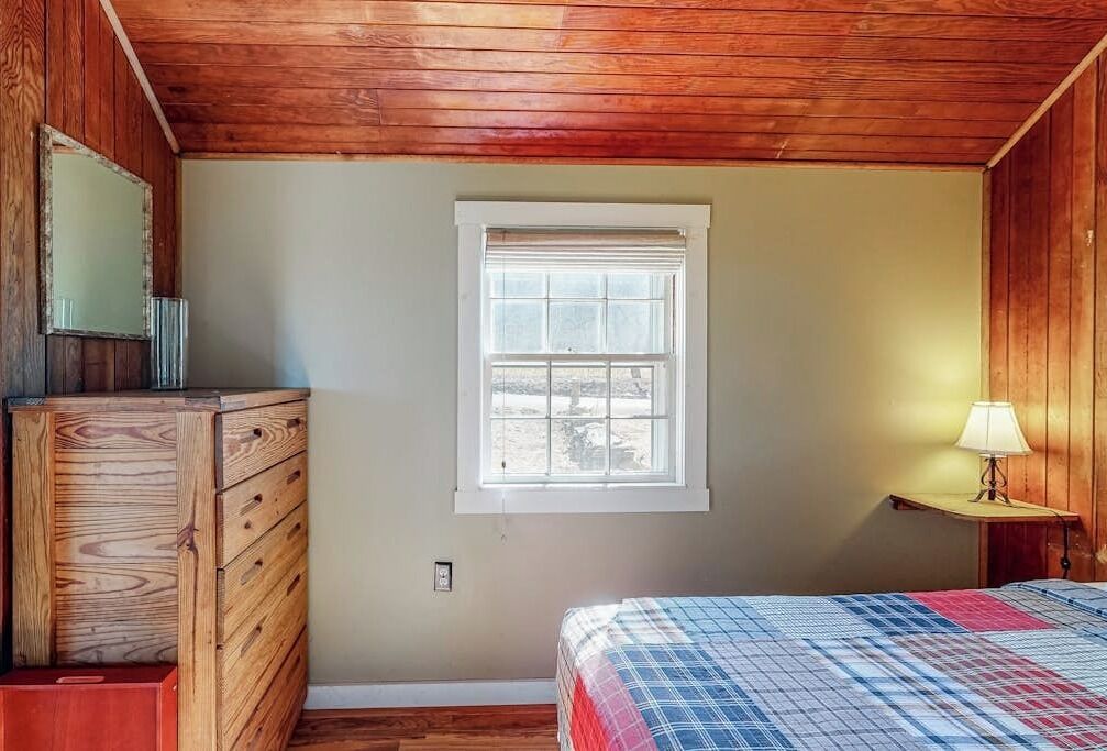 A bedroom with a wooden dresser, a bed with a plaid bedspread, a window, and a nightstand with a lamp.
