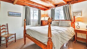 A bedroom with a wooden four-poster bed, two windows with curtains, a bedside table with a lamp, and a chair.