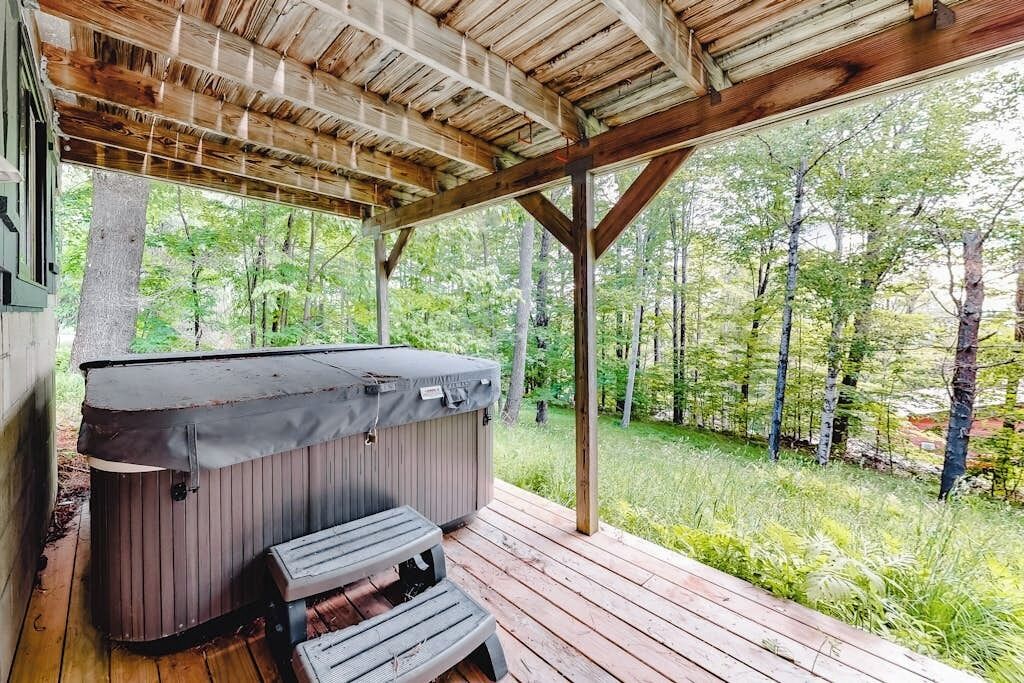 Terraza techada al aire libre con un jacuzzi y escalones que llevan a él, rodeada de zona boscosa.