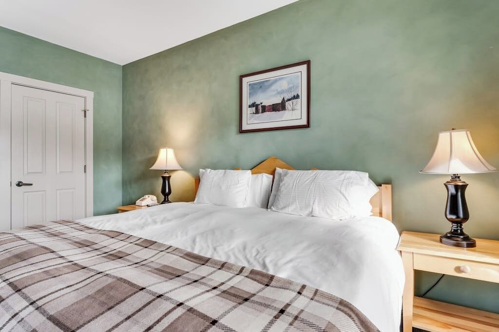 A bedroom with a bed, two bedside lamps, a nightstand, a framed picture on the wall, and a door.