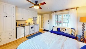 A bedroom with a bed, a window with curtains, a ceiling fan, and a kitchenette.