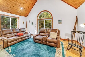 Ein geräumiges Wohnzimmer mit zwei Ledersofas, einer Holzbalkendecke, großen Fenstern und einem rustikalen Sessel.