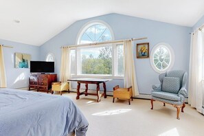 Ein Schlafzimmer mit einem Bett, einem Fernseher, einem Schreibtisch, einem Sessel und großen Fenstern.