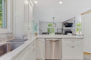 A kitchen with white cabinetry, a stainless steel dishwasher, and a stone fireplace. - Peaceful mountain home near skiing, hiking, mtn biking & more (Jamaica)