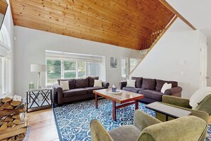 A spacious living room with a high wooden ceiling, a large window, and a staircase leading to another level. - Peaceful mountain home near skiing, hiking, mtn biking & more (Jamaica)