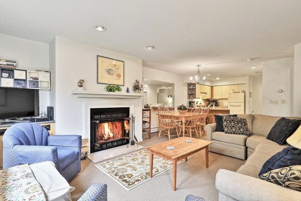 A living room with a fireplace, a television, a dining area, and a kitchen in the background. - Inviting condo with a fireplace, hot tubs, pool, tennis & washer/dryer (Ludlow)