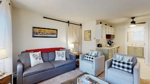 Een woonkamer met een bank, fauteuils, een salontafel en een keuken op de achtergrond.
