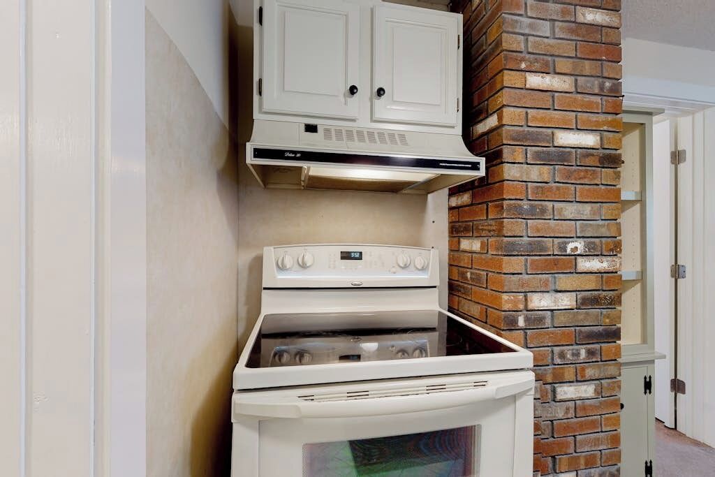 Una cocina con estufa blanca, pared de ladrillo y un armario blanco arriba.