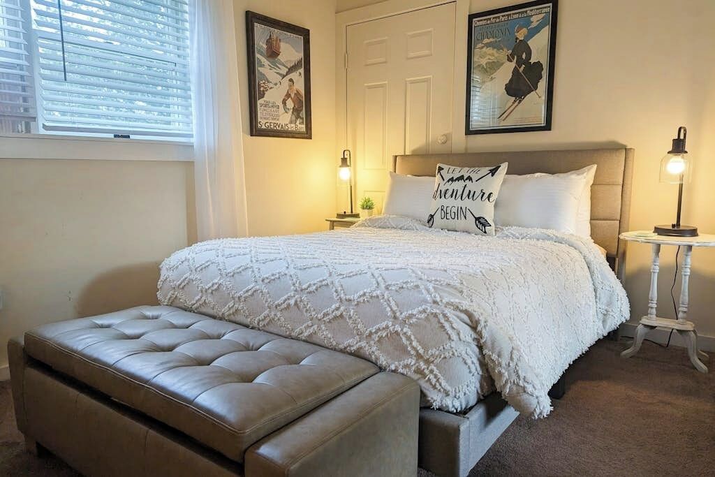 A bedroom with a bed, a bedside table, a lamp, a bench, and two wall-mounted pictures.