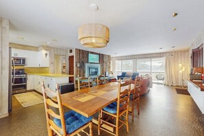 A spacious living area with a wooden dining table, chairs, a large window, and a kitchen in the background.