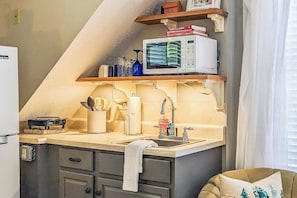 Eine kleine Küche mit Mikrowelle, Spüle und einer kompakten Arbeitsplatte. Darüber befinden sich Holzregale mit Büchern und einem kleinen Hausmodell.