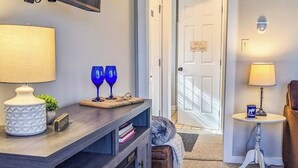 A living room with a dark wooden console table, a white lamp, two blue wine glasses, a small plant, and a door with a sign. - Connecting condos w/ electric fireplace, near Okemo mountain, wifi (Ludlow)