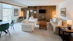 Een woonkamer met een houten wandpaneel, een flatscreentelevisie en een eethoek met groene stoelen.