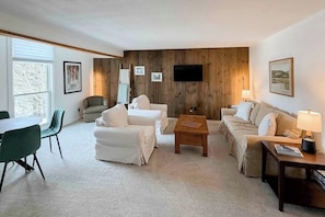 A living room with a wooden wall panel, a flat-screen TV, and a dining area with green chairs. - Skiers apartment, walk to lifts at Sundance Resort, mountain views, shuttle, w/d (Dover)