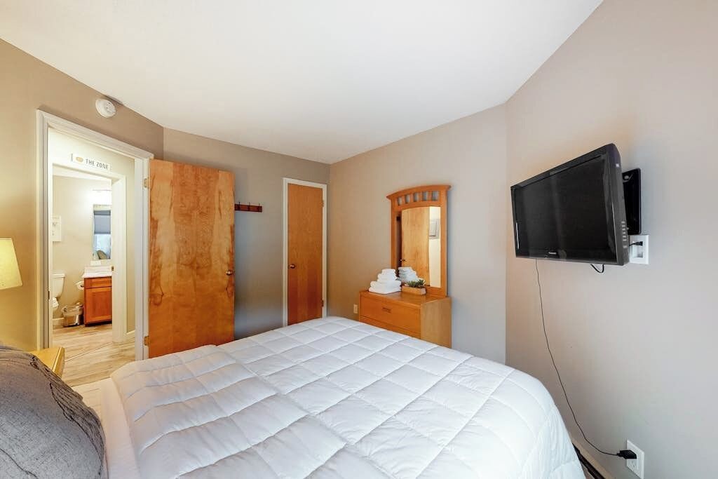 Un dormitorio con cama, televisor, cómoda, espejo y puerta que da a un baño.