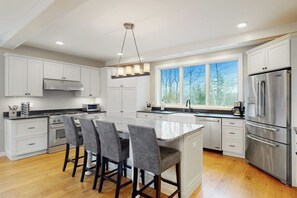 A modern kitchen with white cabinetry, a large island with bar stools, stainless steel appliances, and a view through the windows. - Ski-in/out home with a hot tub, fireplace, cable & golf nearby (Ludlow)