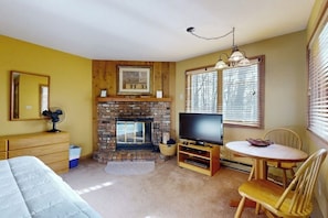 A room with a fireplace, a television, a bed, a dining table with chairs, and a mirror. - Ski-in/out Condo on Bull Run Trail with fireplace, & full kitchen (Ludlow)