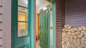 Une porte verte avec une fenêtre, une pile de bois de chauffage et un espace intérieur visible, éclairé par un luminaire.