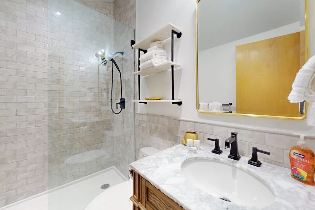 A bathroom with a glass shower enclosure, a white sink with a marble countertop, and a large mirror with a gold frame.