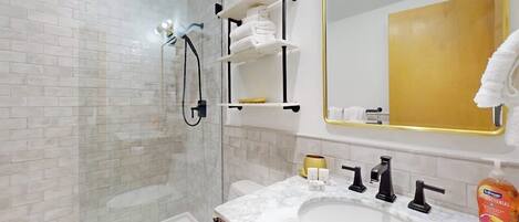 A bathroom with a glass shower enclosure, a white sink with a marble countertop, and a large mirror with a gold frame.