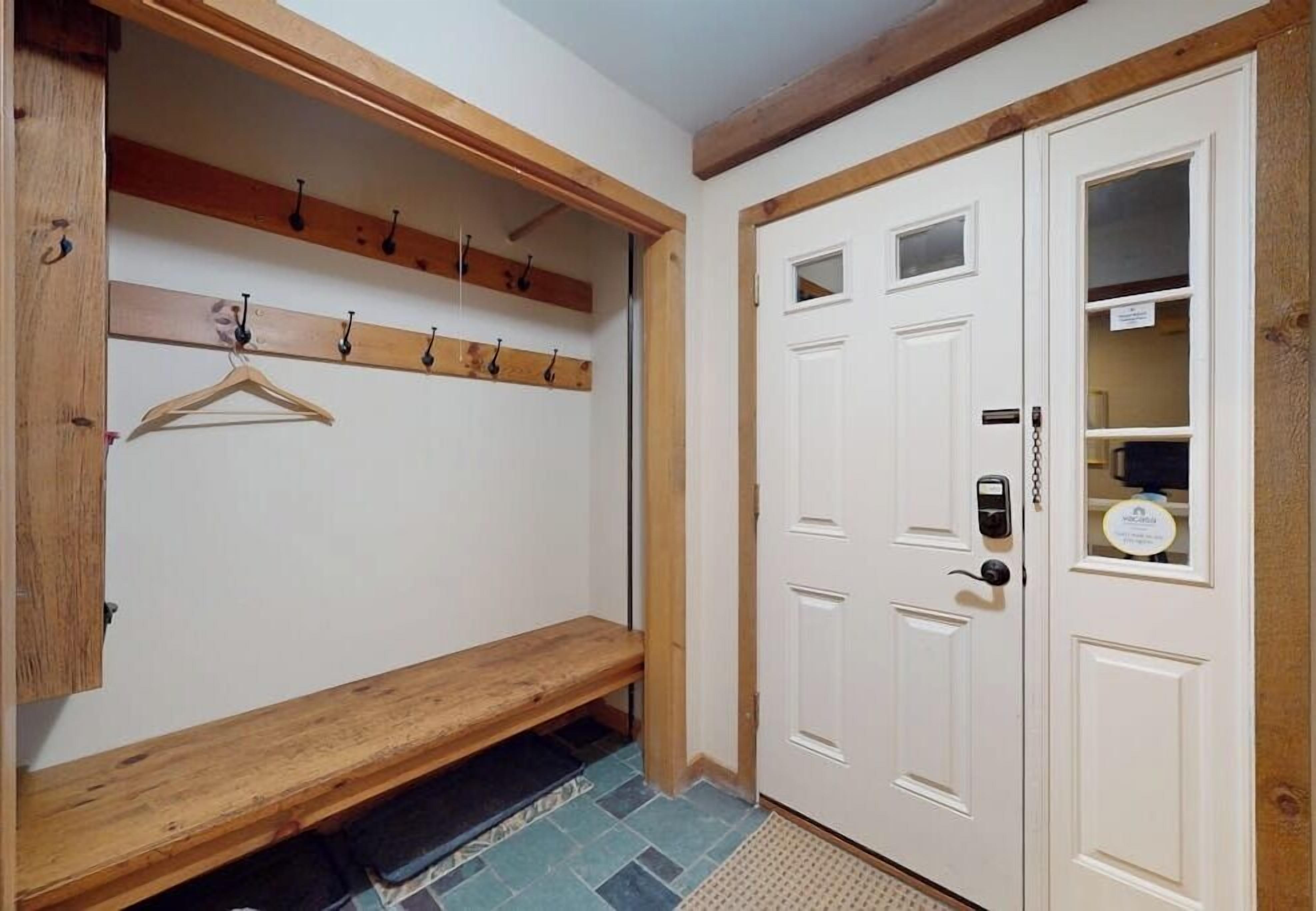 Um corredor com um banco, ganchos para casacos, uma porta com olho mágico e uma pequena prateleira com uma placa.