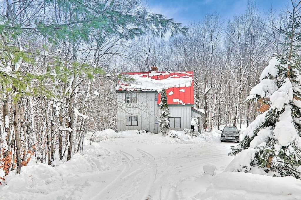 A snow-covered house with a red roof, surrounded by trees and a snow-covered driveway.