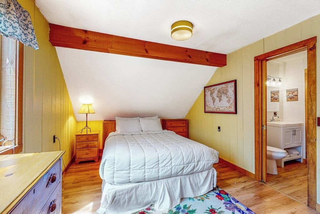 A bedroom with a bed, wooden floor, a nightstand, a lamp, a door leading to a bathroom, and a ceiling with wooden beams.