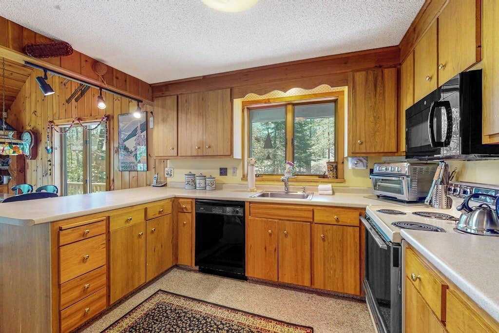 A wooden kitchen with a dishwasher, stove, microwave, and a window with a view of trees.