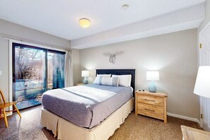 A bedroom with a bed, two bedside tables with lamps, a chair, a sliding glass door to a balcony, and a wall-mounted decor piece. - Winterplace ground-floor ski-in/ski-out condo on the Lift Line trail (Ludlow)