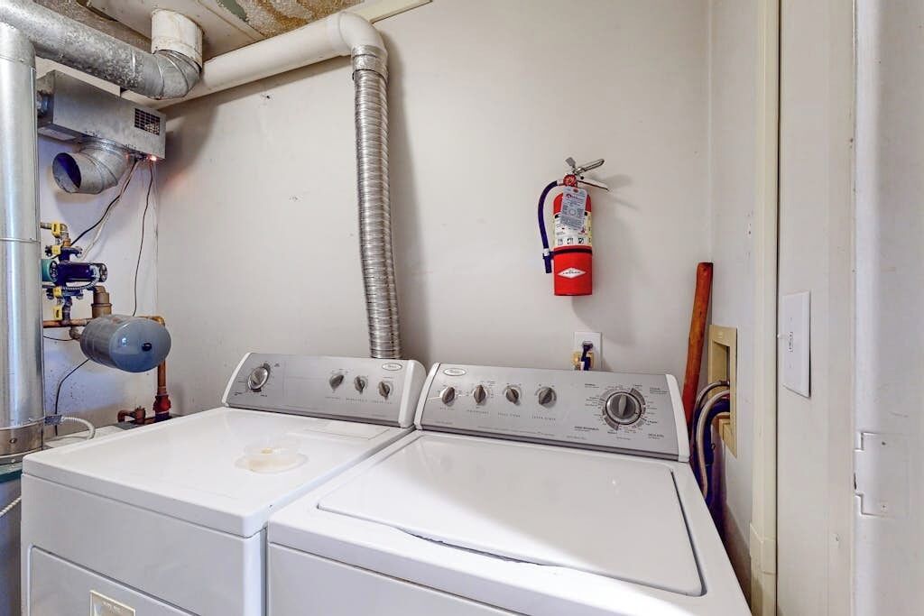 A laundry room with two stacked washers and dryers, a fire extinguisher on the wall, and visible plumbing and ductwork.
