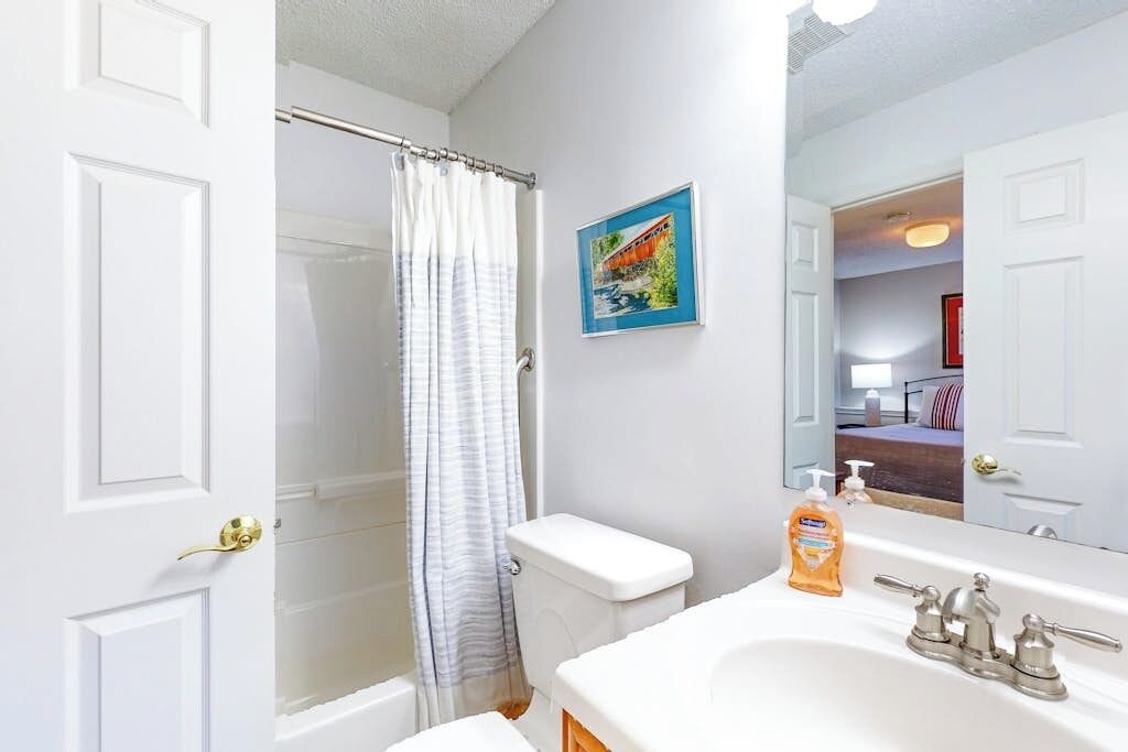 A bathroom with a shower curtain, toilet, sink, and a mirror reflecting a bedroom.