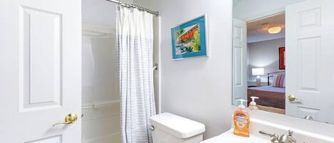 A bathroom with a shower curtain, toilet, sink, and a mirror reflecting a bedroom.