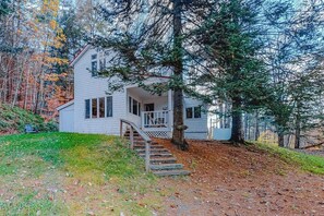 A white house with a porch and a staircase leading up to it, surrounded by trees and a grassy area. - Spacious Home w/Seasonal Lake View, Washer/Dryer & Cable (Ludlow)