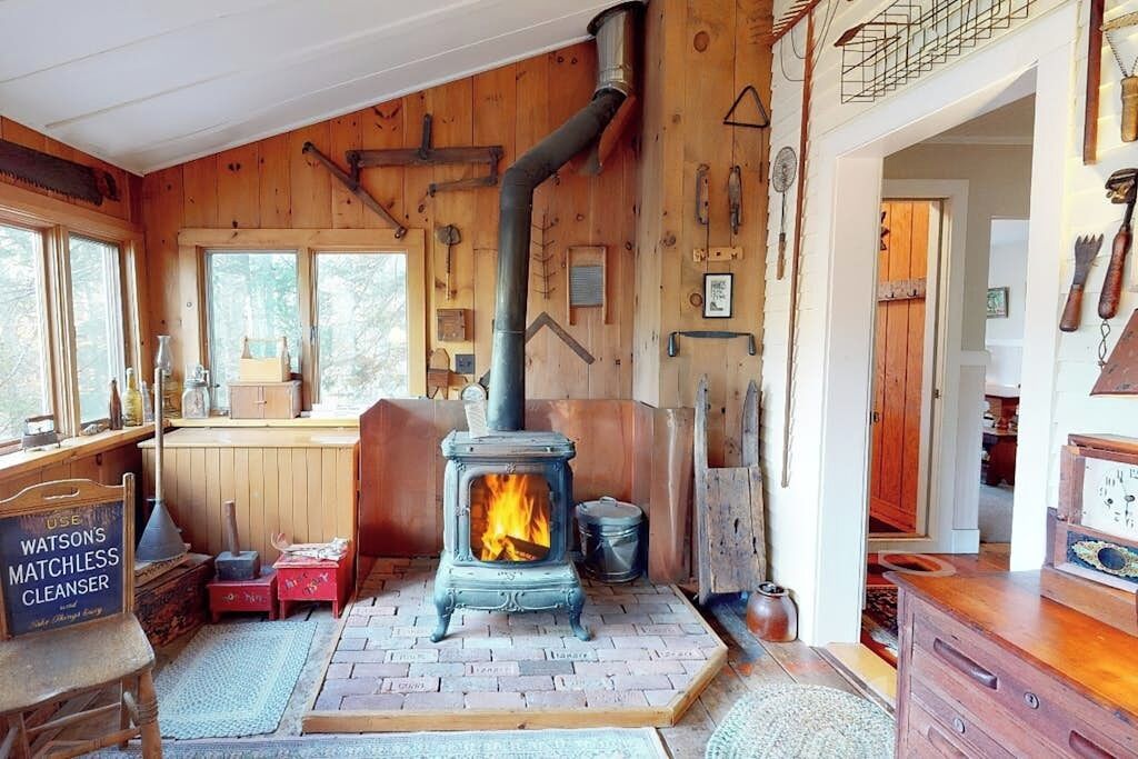 A rustic cabin interior with a wood-burning stove, wooden walls, a window, and a wooden bench.