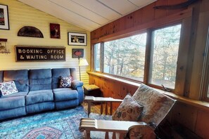 A cozy cabin interior with a blue sofa, a wooden chair, a rug, and a window with a view of trees. - Spacious Home w/Seasonal Lake View, Washer/Dryer & Cable (Ludlow)