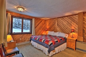 Ein gemütliches Schlafzimmer mit einer holzgetäfelten Wand, einem großen Bett mit farbenfroher Steppdecke, einem Fenster mit Blick auf schneebedeckte Bäume und zwei Nachttischlampen.