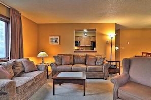 Ein Wohnzimmer mit Sofa, Sessel, Couchtisch und Lampe. Im Hintergrund ist eine Küchenzeile erkennbar.