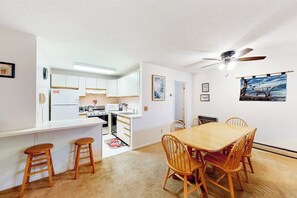 Cocina compacta con armarios blancos, comedor con sillas de madera y ventilador de techo.