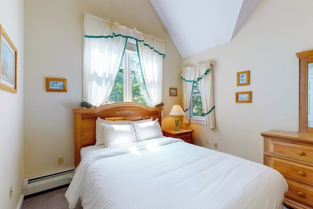 Un dormitorio con cama, mesita de noche, cómoda y dos ventanas con cortinas.