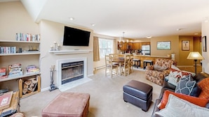 A spacious living room with a fireplace, a television, a dining area, and a kitchen. - Condo near ski trail with fireplace, hot tub, pool, tennis & washer/dryer (Ludlow)