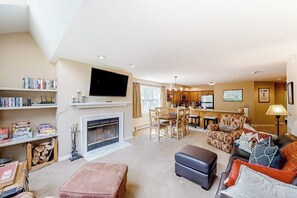 A spacious living room with a fireplace, a television, a dining area, and a kitchen. - Condo near ski trail with fireplace, hot tub, pool, tennis & washer/dryer (Ludlow)