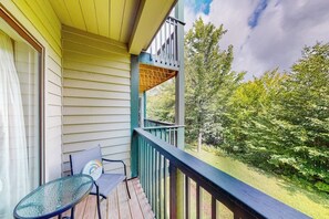 A wooden deck with a glass table and chairs, a balcony with a railing, and a view of trees. - Condo near ski trail with fireplace, hot tub, pool, tennis & washer/dryer (Ludlow)