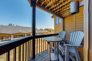 Eine Holzterrasse mit zwei Adirondack-Stühlen und einem kleinen runden Tisch, mit Blick auf ein Gebäude mit einem geneigten Dach.