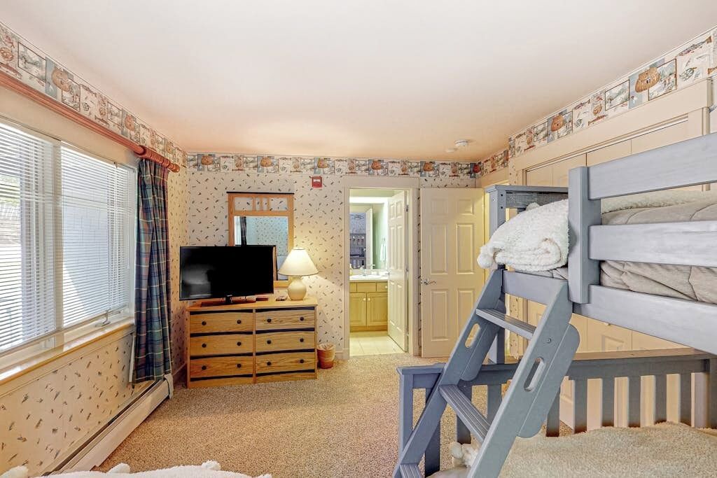 Une chambre avec un lit superposé, un téléviseur posé sur une commode en bois, une lampe et une fenêtre avec des stores.