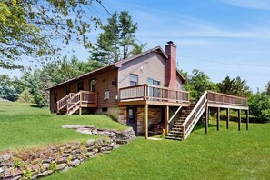 A two-story house with a stone foundation, wooden deck, and a staircase leading to a balcony. - Quiet country home w/ large deck & fireplace, near Okemo Mtn & town (Ludlow)